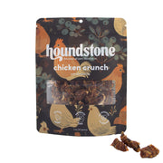 A package of Houndstone Chicken Crunch dehydrated dog treats on a white background, with three loose pieces of dog treats shown in the bottom right corner. The packaging is dark brown with illustrations of chickens and blueberries, and a clear window in the center reveals the textured dog treat pieces inside.