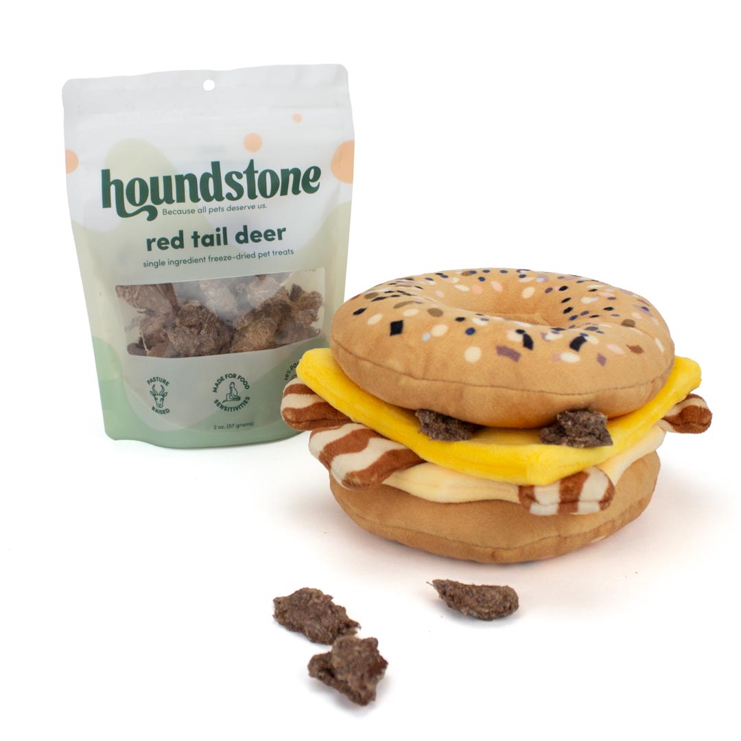 Houndstone Red Tail Deer single ingredient freeze dried dog treats shown next to a novelty plush toy shaped like a bagel cheese burger, with a few loose dog treats in front on a white background.