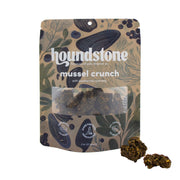 A stand-up pouch of Houndstone Mussel Crunch dehydrated dog treats on a white background, with two pieces of the dehydrated dog treats placed in front of the package.